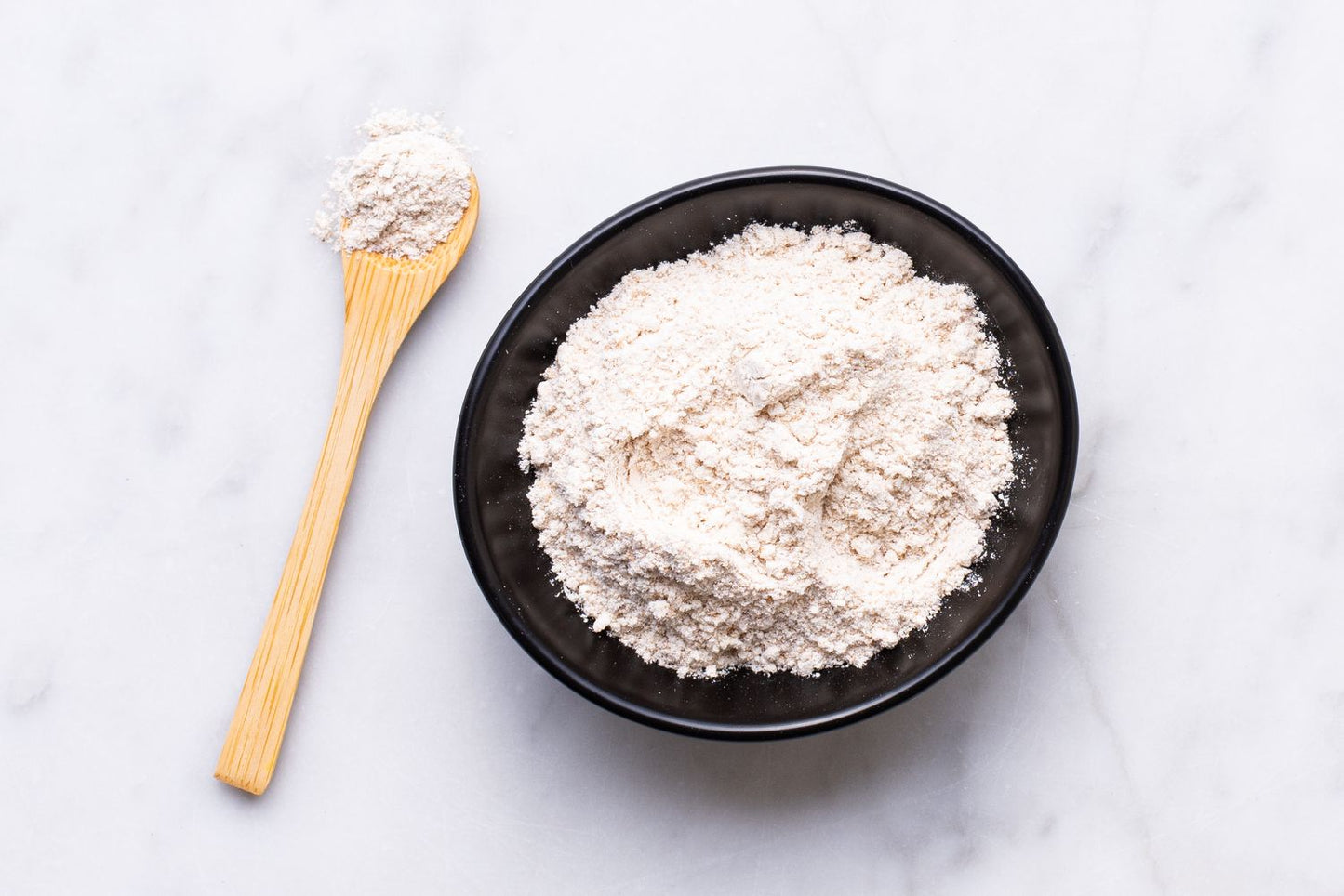A black bowl filled with white powdery substance sits on a white marble surface, accompanied by a wooden spoon.
