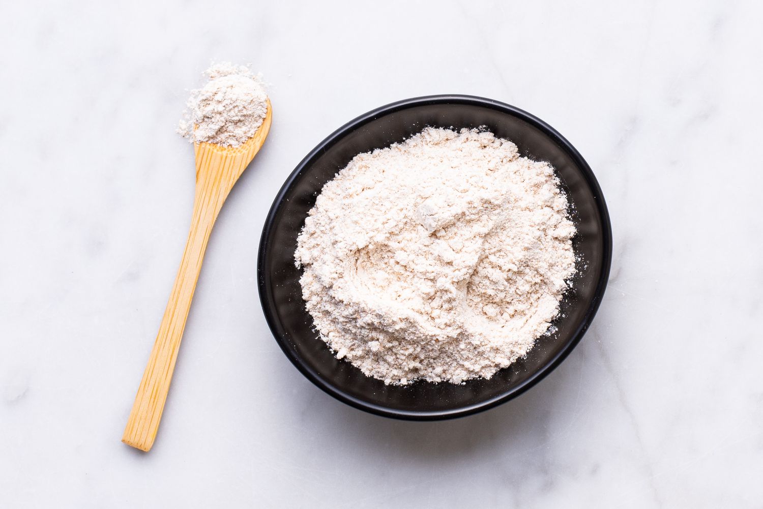A black bowl filled with white powdery substance sits on a white marble surface, accompanied by a wooden spoon.