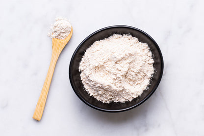 A black bowl filled with white powdery substance sits on a white marble surface, accompanied by a wooden spoon.