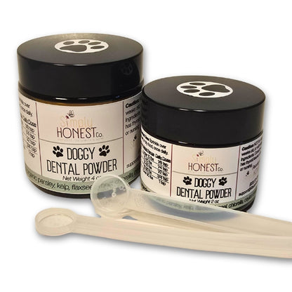 Two jars of "DOGGY DENTAL POWDER" with a black lid and a clear plastic scoop, along with a clear plastic spoon.